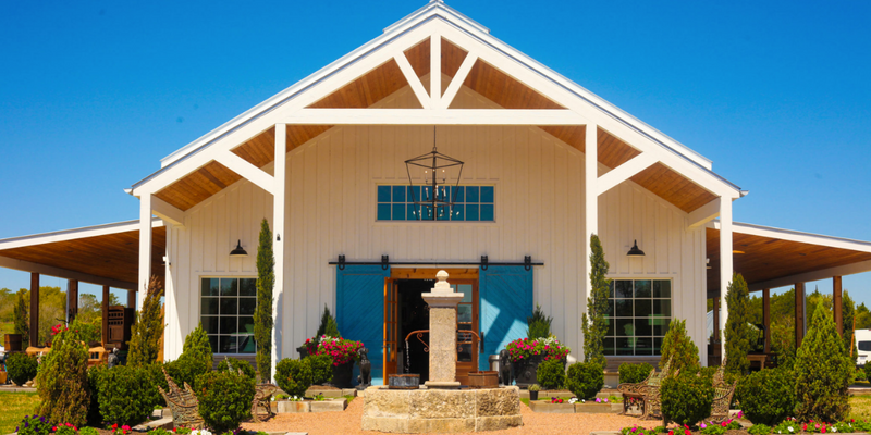 Entrance to an antique barn at The Compound in Round Top, Texas, with a white gabled roof, hanging chandelier, and a fountain in the courtyard