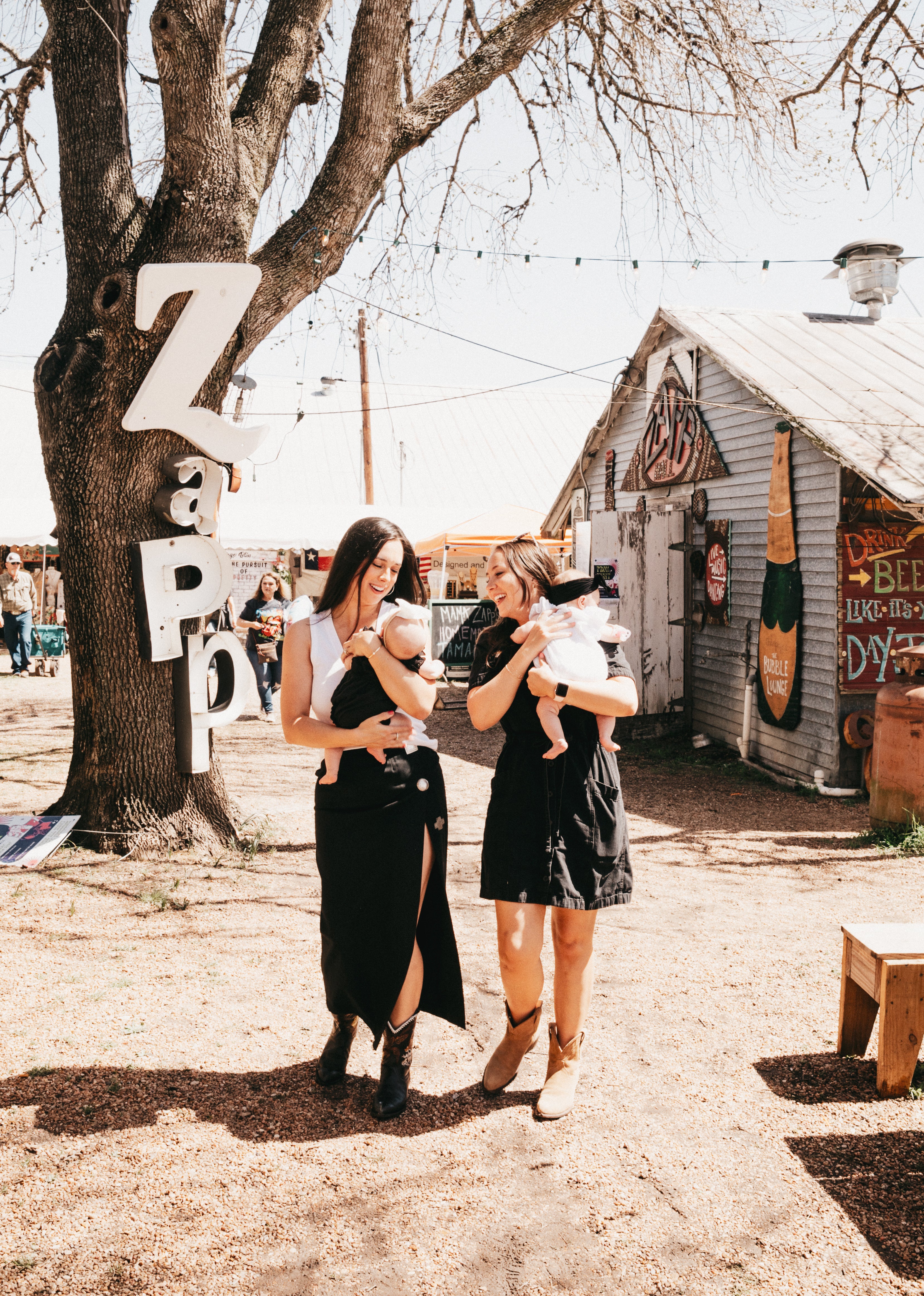 Two friends, one holding a baby, standing in front of large white letters spelling ZAPP at Zapp Hall in Round Top Texas