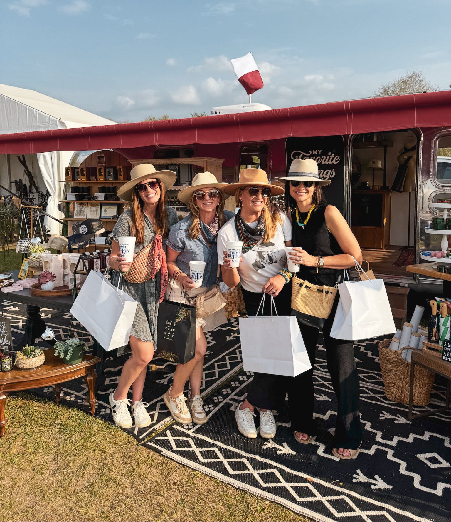 Three friends holding shopping bags and drinks posing under a Texas flag at a vendor booth during the Round Top antique show