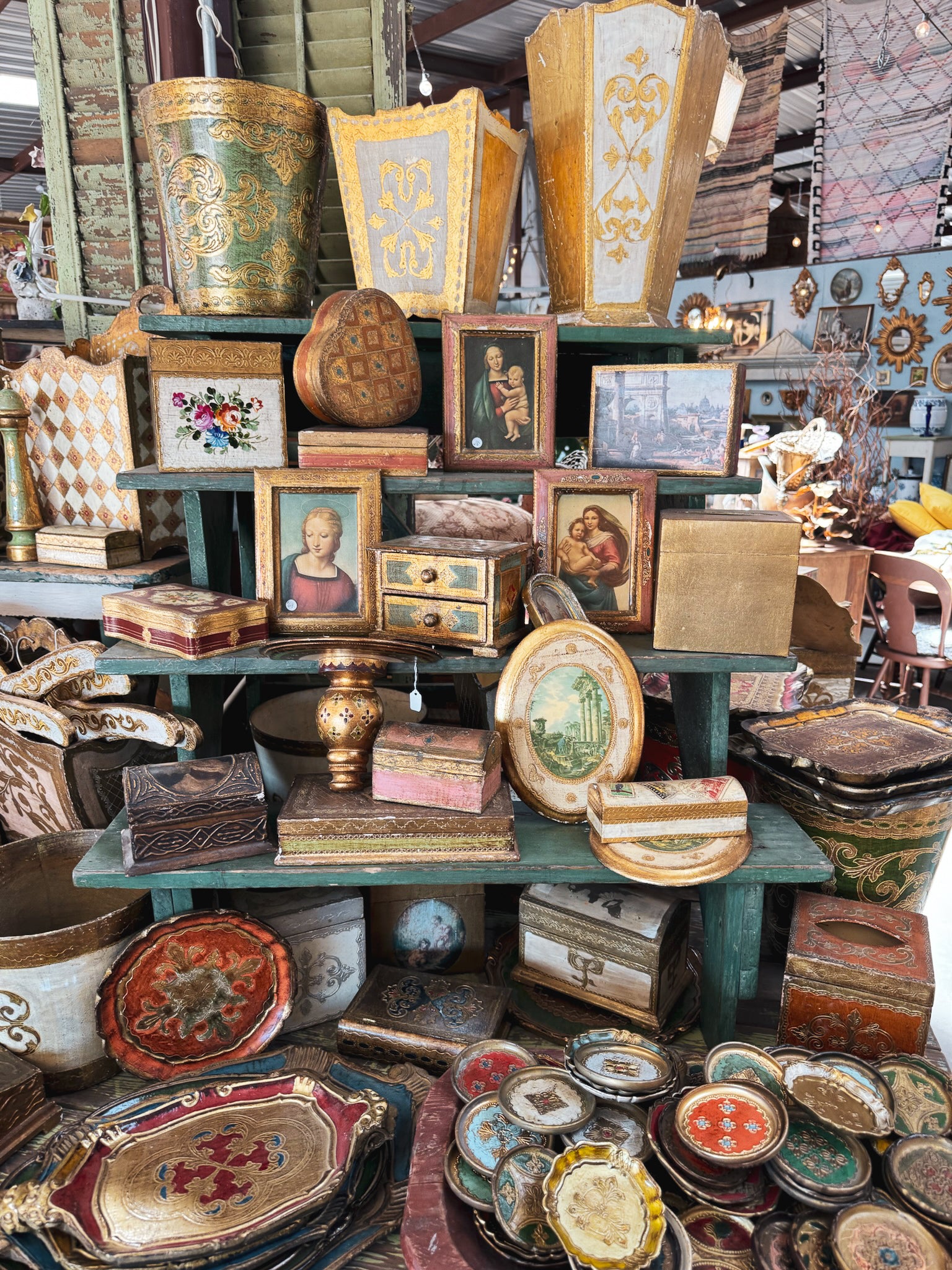 A colorful collection of vintage framed art, religious paintings, gilded decorative boxes, and antique home goods stacked at a Round Top vendor booth